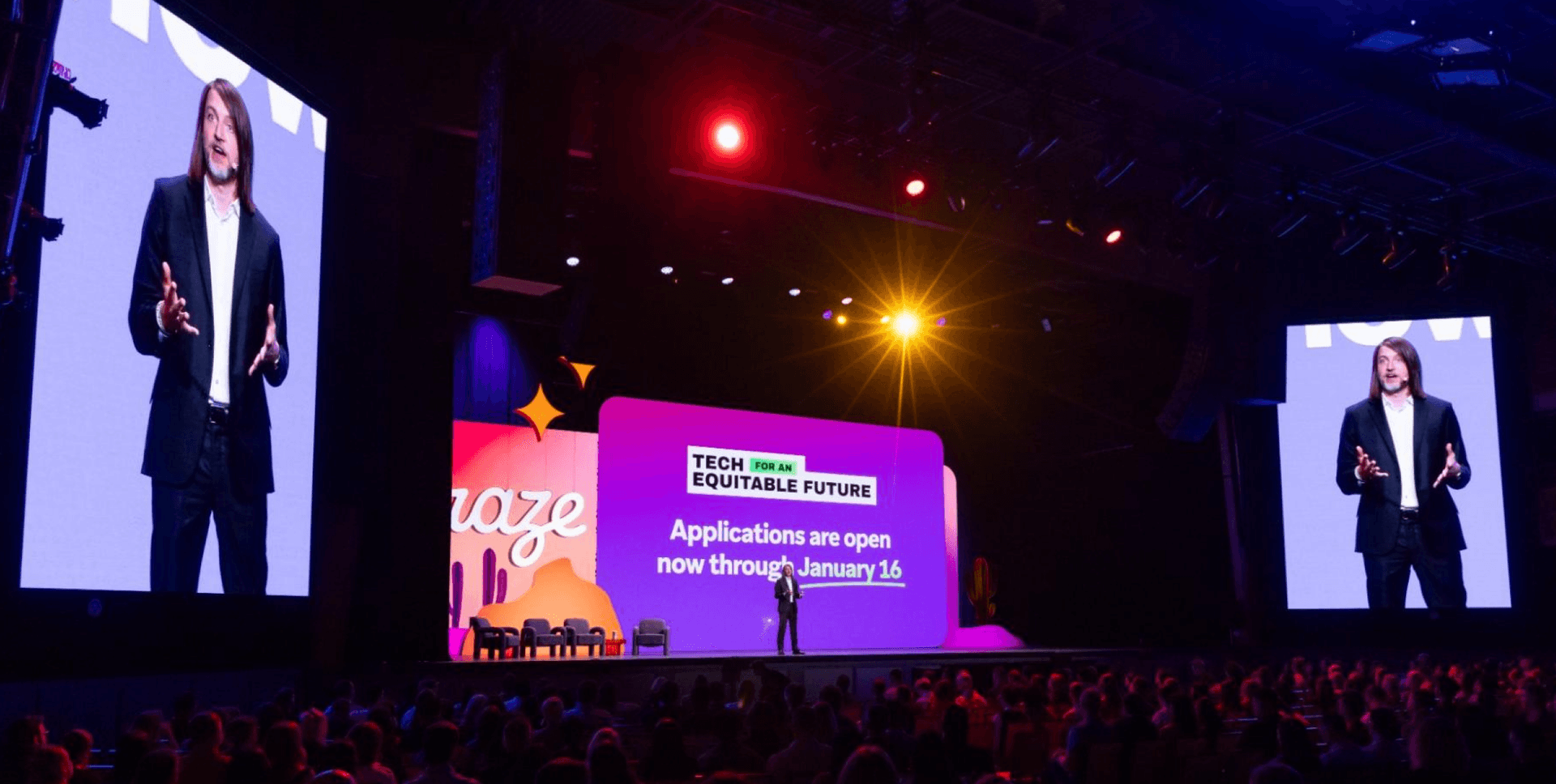 A speaker on stage addresses an audience at a large conference, with screens displaying the speaker and event branding.