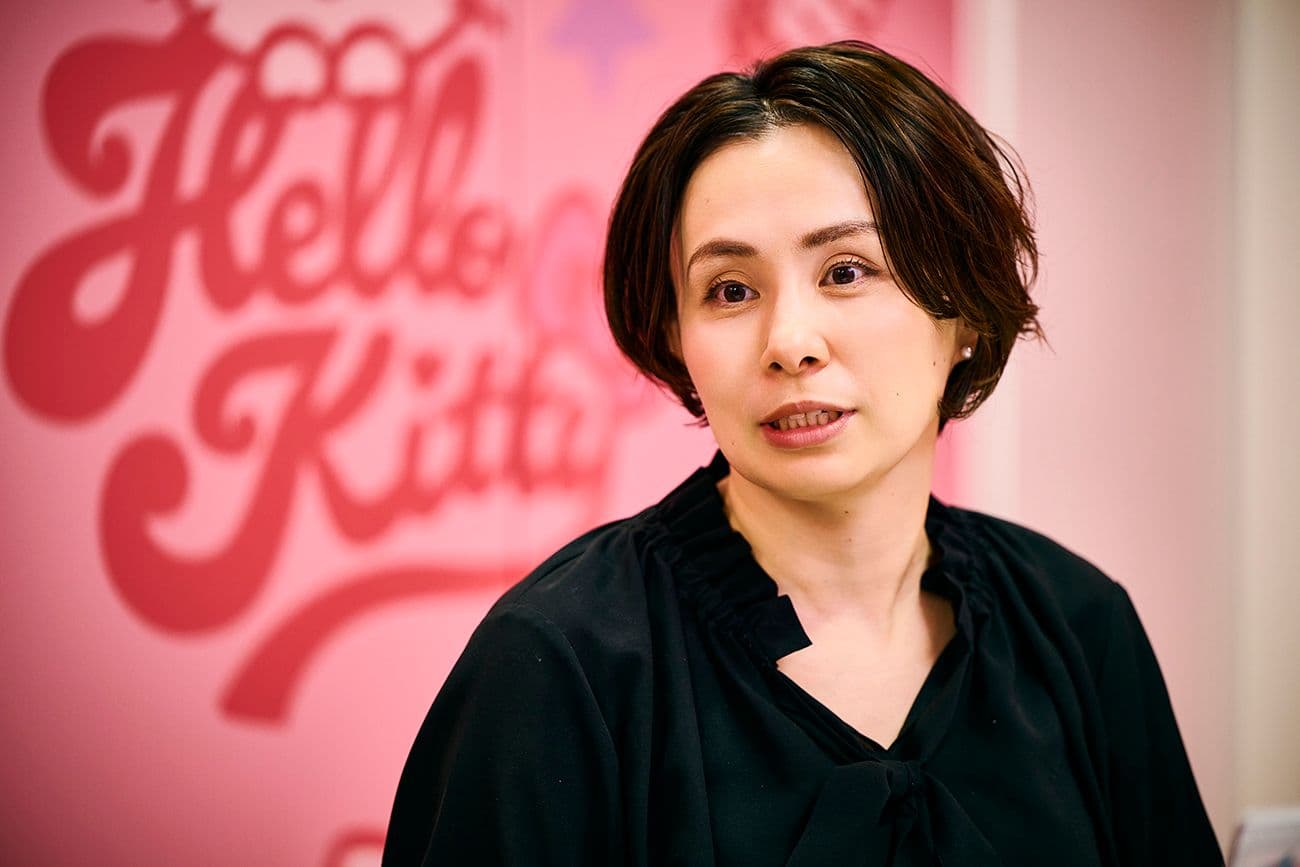 ピンクの壁を背に、ぼやけたハローキティのロゴを見ながら話す女性(サンリオ デジタル事業本部 デジタル事業開発部 CX推進課 シニアマネージャー 鈴木 理恵氏)。