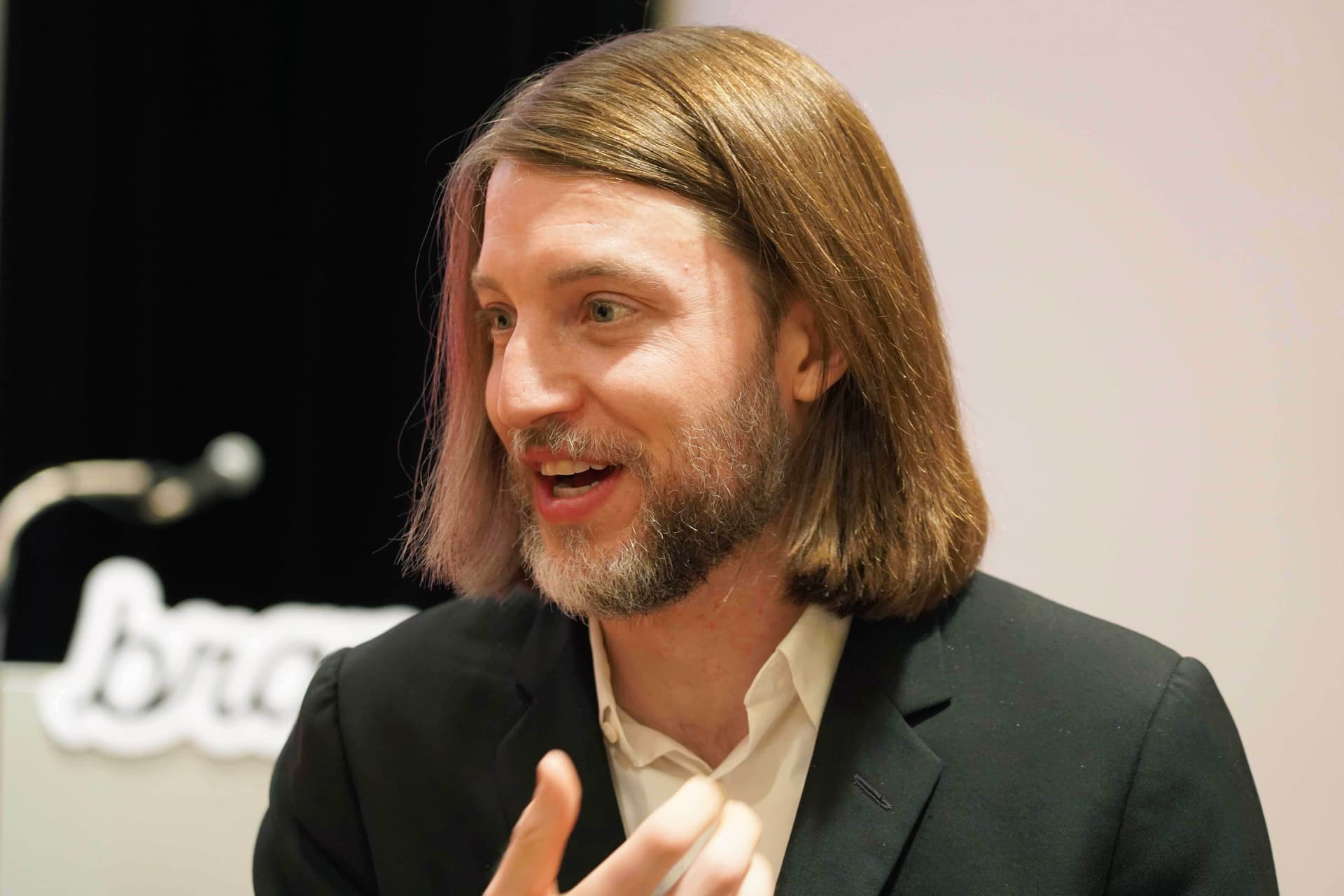 a man with long hair and a beard is wearing a suit and talking into a microphone .