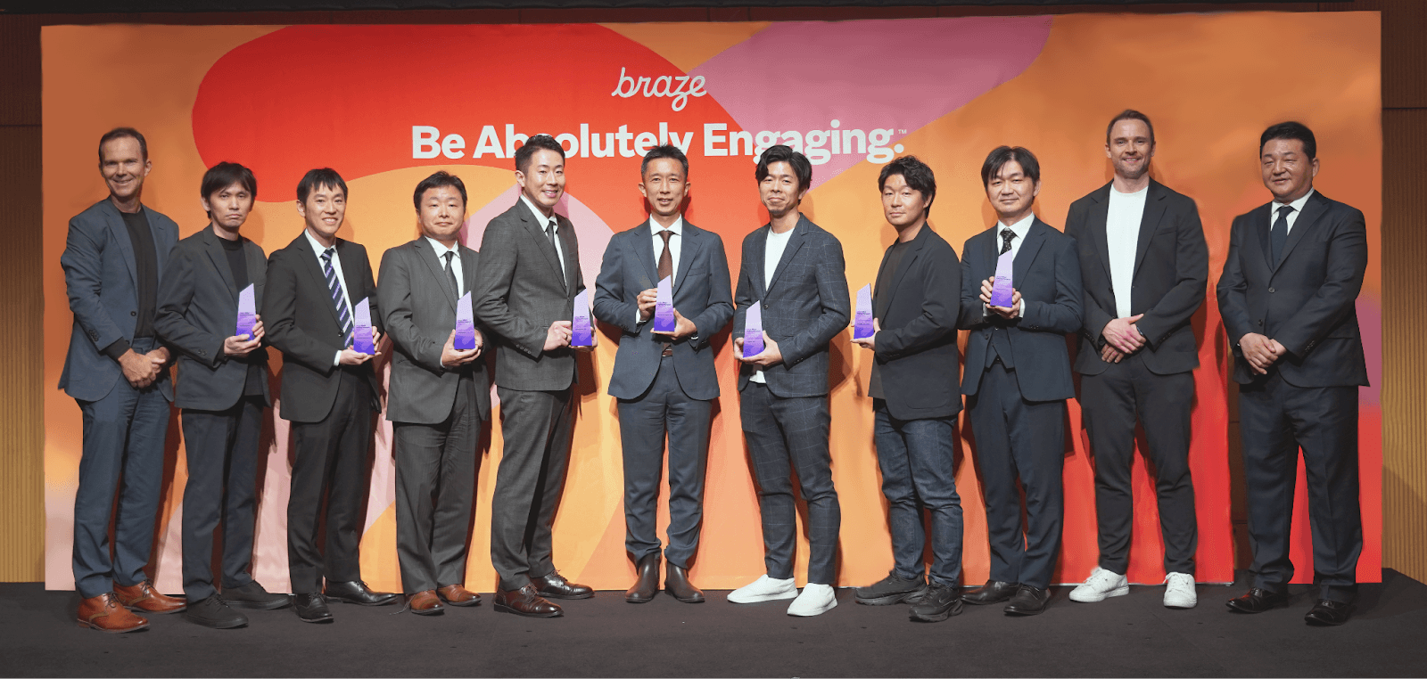 Eleven men, some holding awards, stand on a stage in front of a "Braze: Be Absolutely Engaging" backdrop.