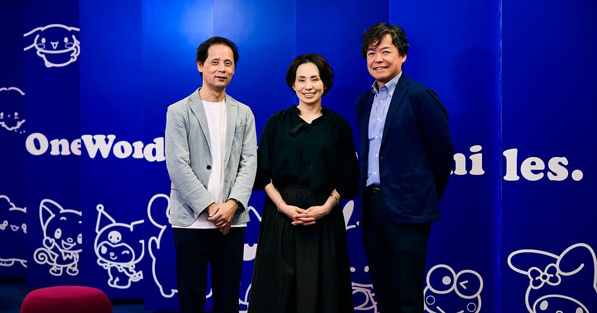 青い壁の前で、二人の男性と一人の女性が笑顔で立っている。壁には漫画のキャラクターのイラストが描かれている。