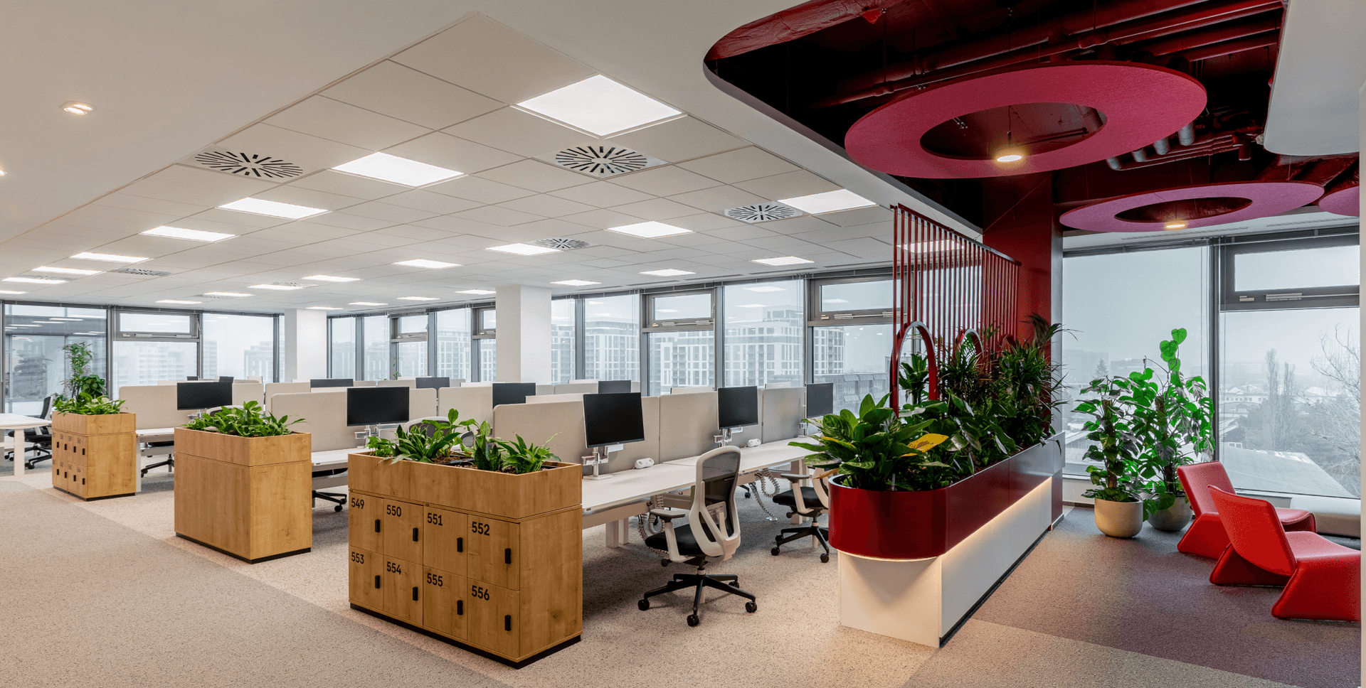 Modern open-plan office with desks, plants, large windows, and a lounge area with red accents.