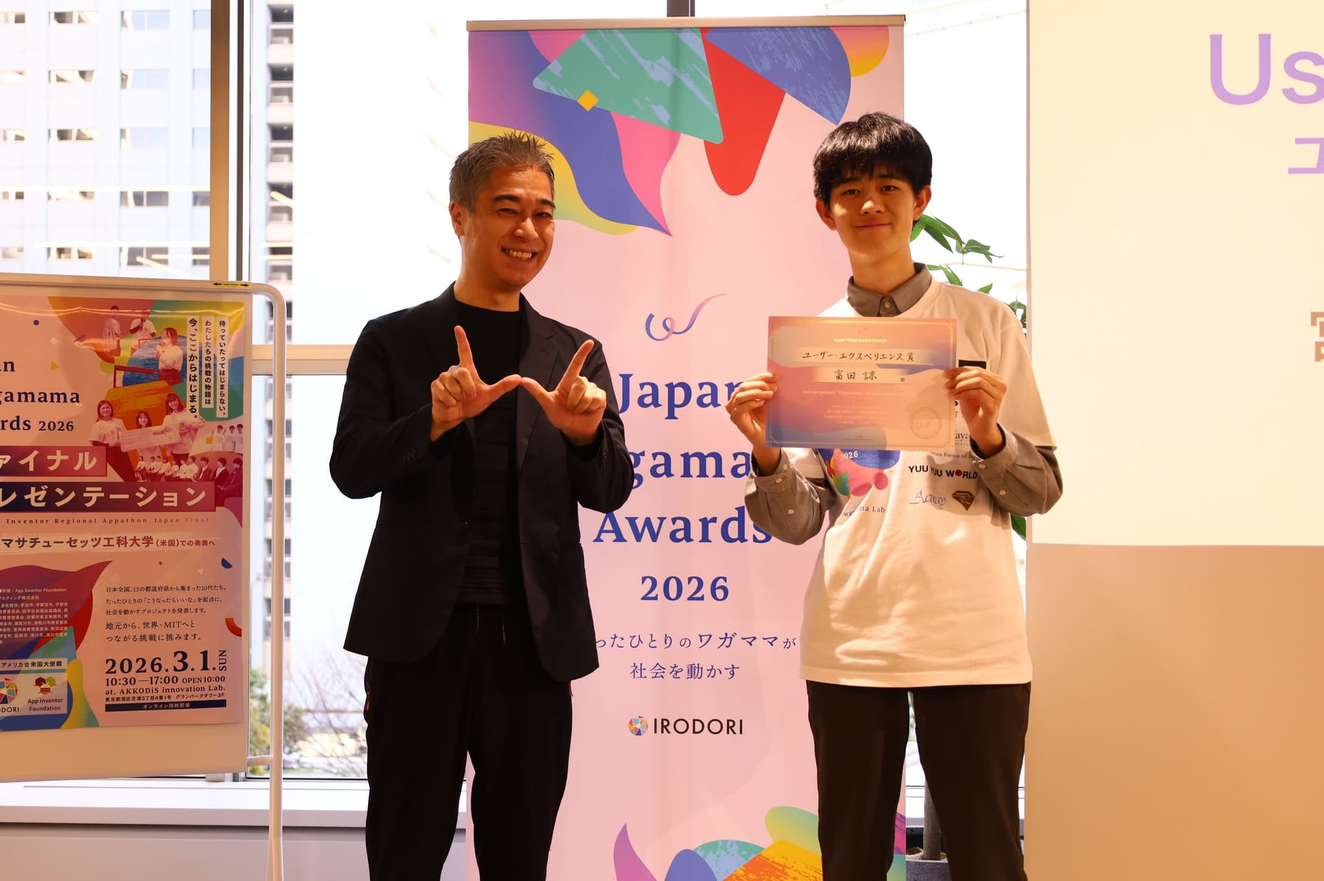 表彰式に出席した二人の男性:一人は笑顔で身振り手振りし、もう一人は表彰状を手にしている。