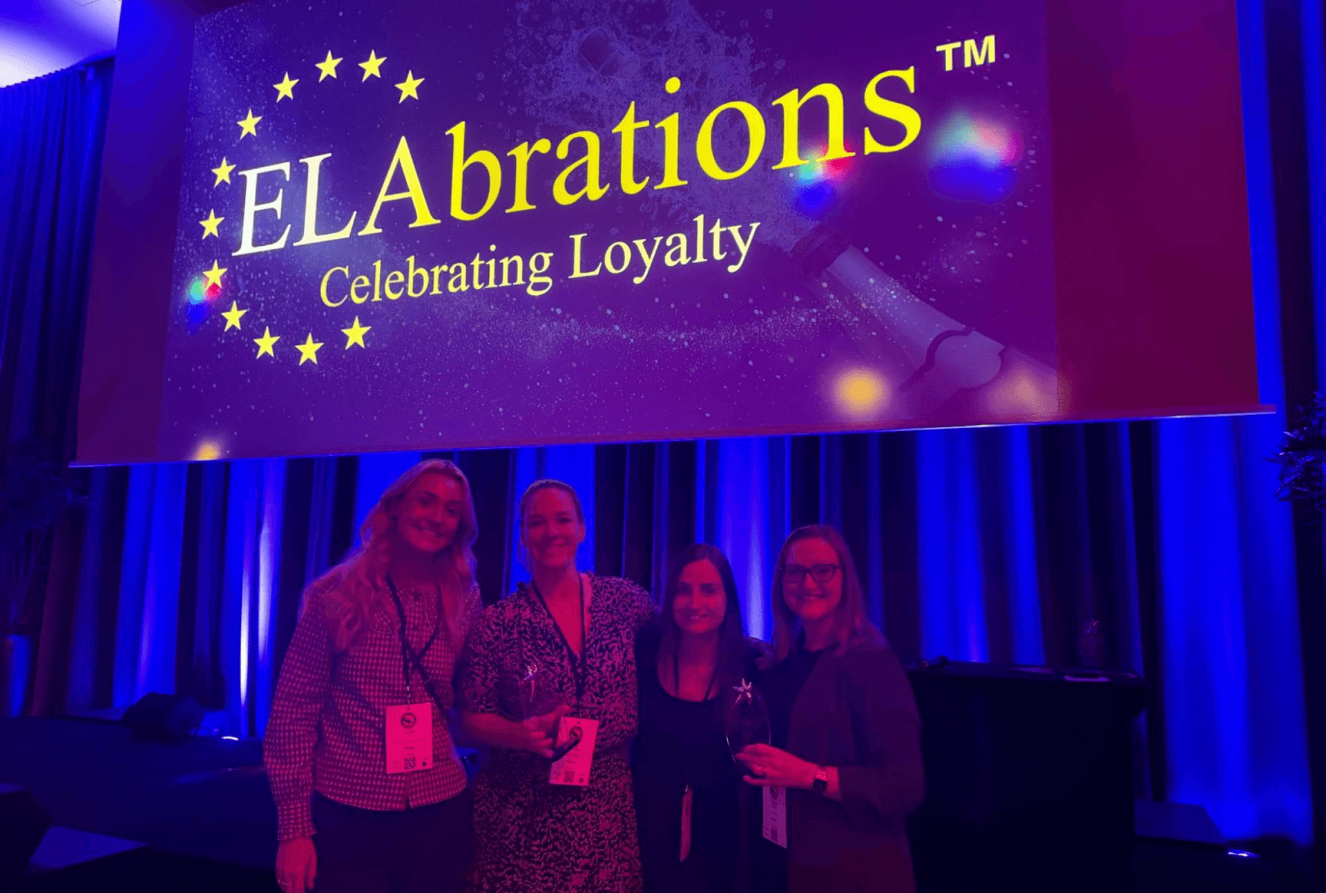 Four smiling women hold awards in front of a screen displaying "ELAbrations Celebrating Loyalty."