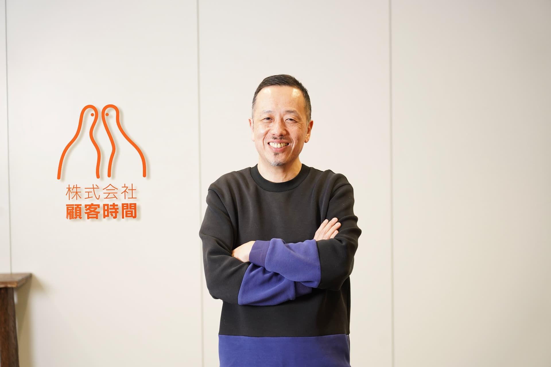腕を組んだ笑顔の男性が、白い壁に描かれたオレンジ色のロゴと日本語の文字の前に立っている。