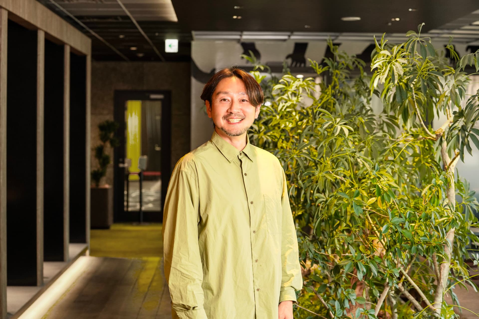 Smiling man in a light green shirt in a modern office setting with plants.
