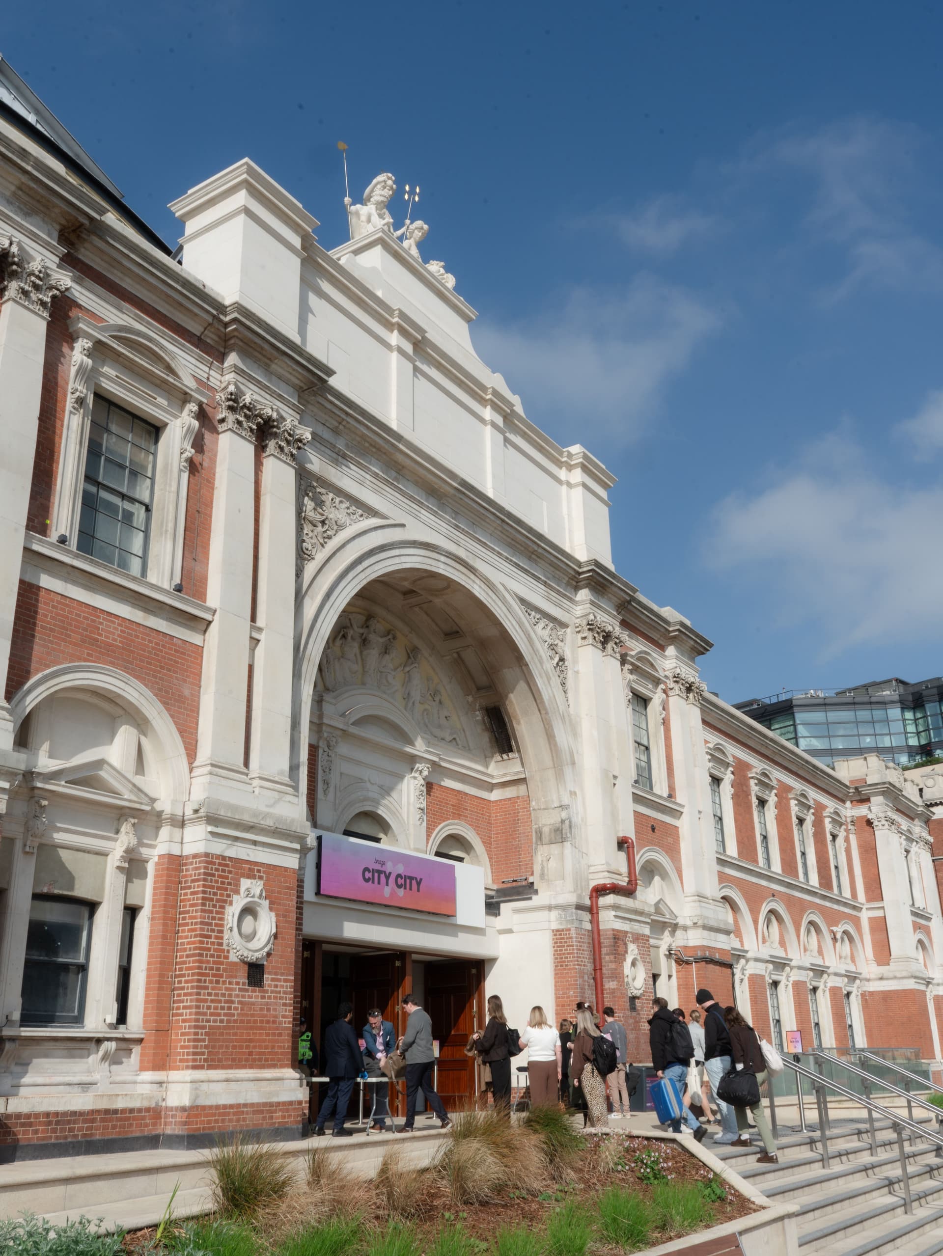 An ornate brick and stone building with an arched entrance and a "THE CITY CITY" sign, with people entering under a blue sky.