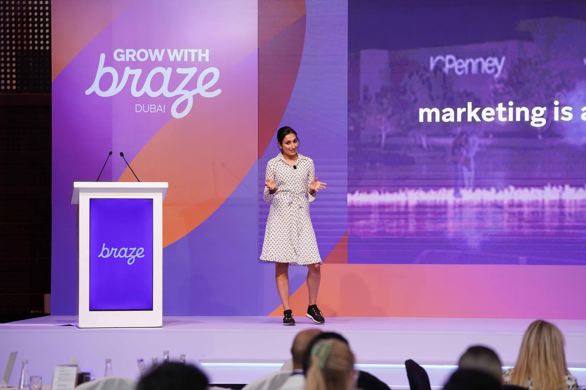 A woman presents on stage at the "Grow with Braze Dubai" event, with a screen showing "JCPenney marketing is a..." in the background.