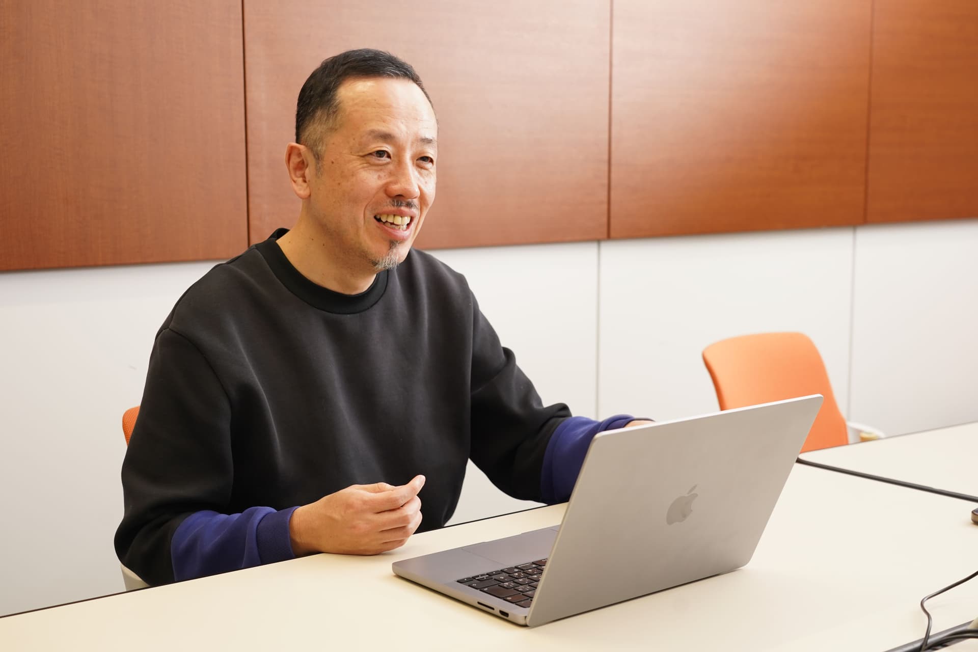 黒いシャツを着た笑顔の男性が、開いたノートパソコンを前にテーブルに座っている。