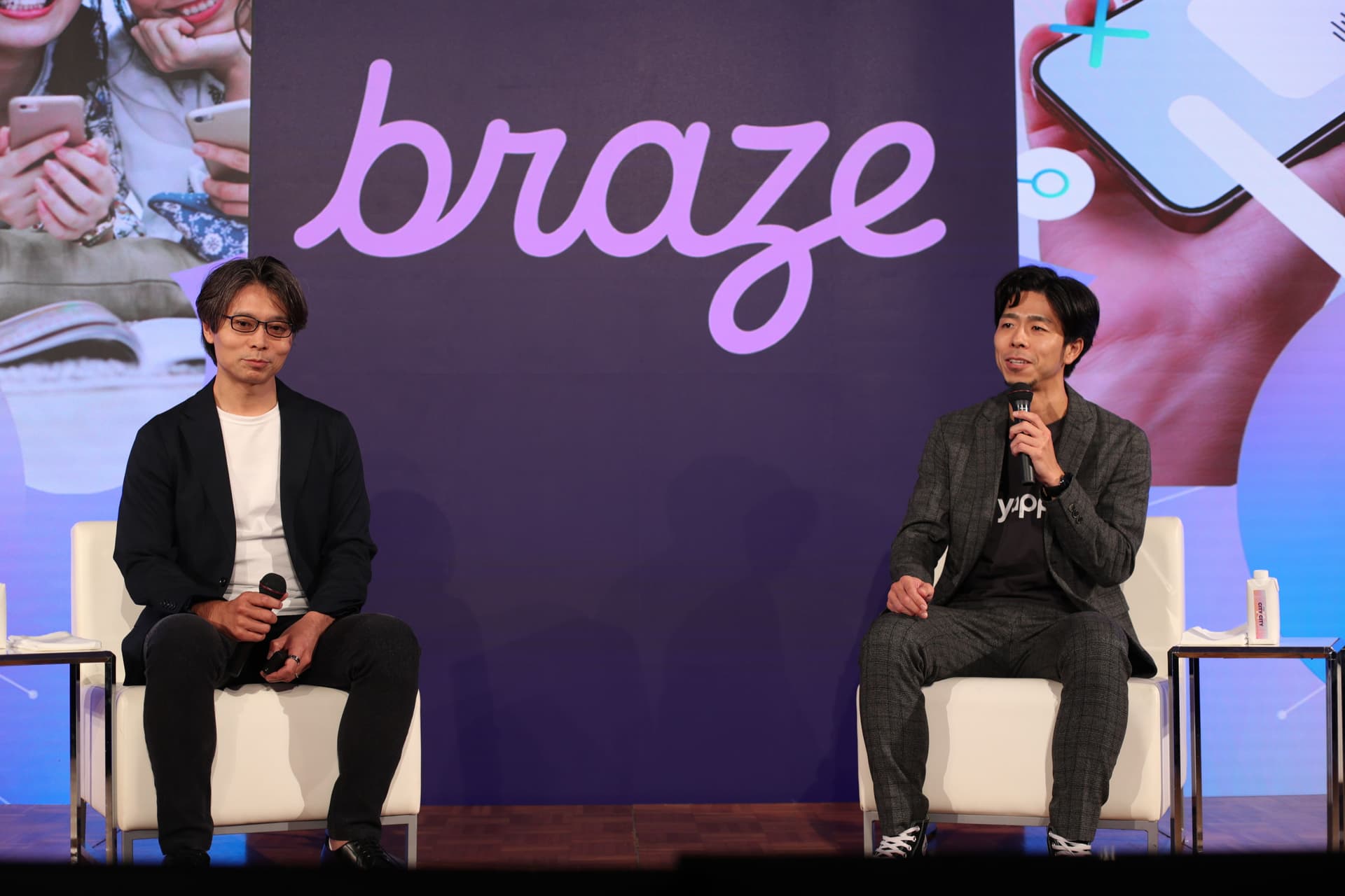 Two men sit on a stage with a purple Braze backdrop, holding microphones.
