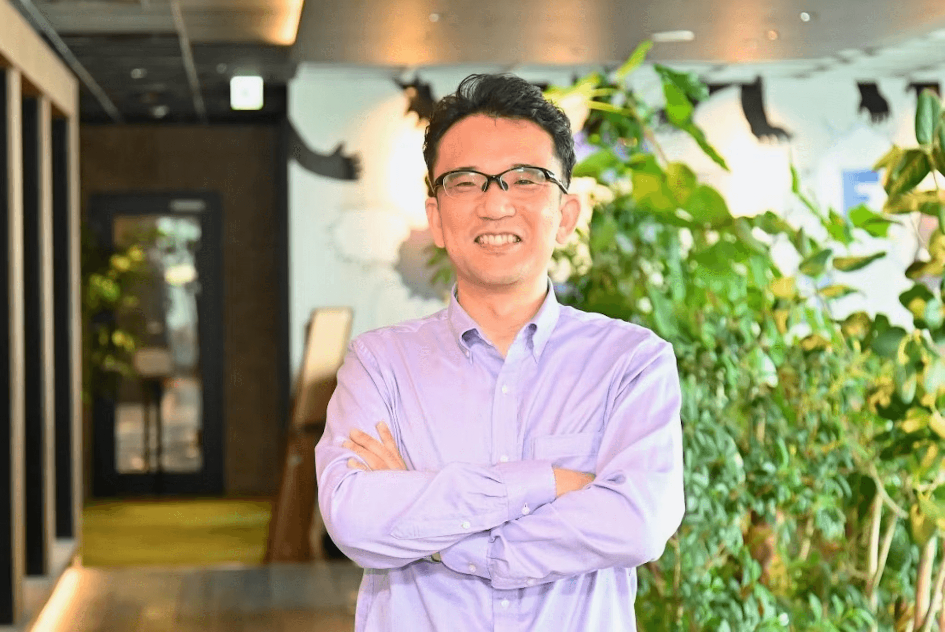 a man wearing glasses and a purple shirt stands with his arms crossed