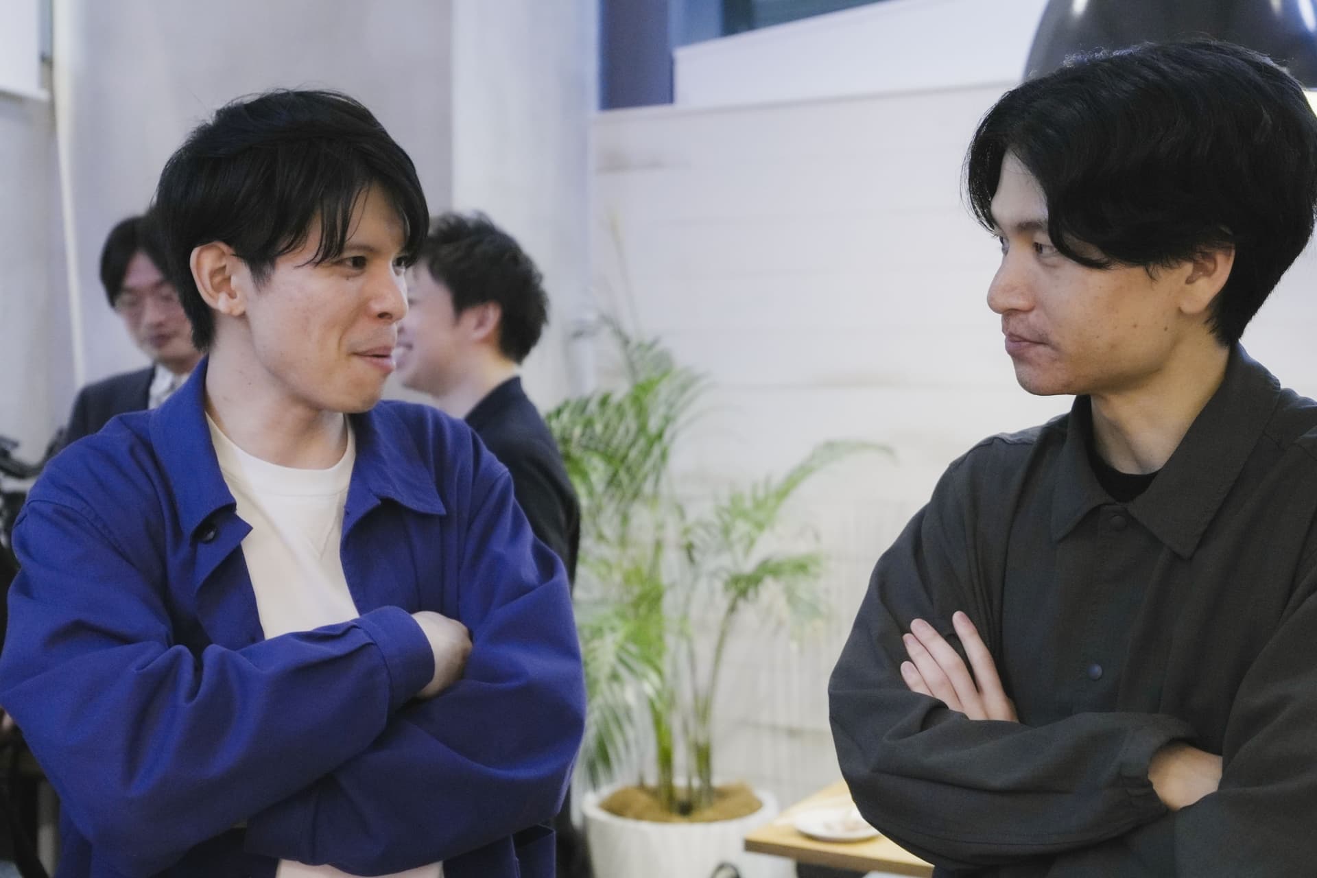 two men are standing next to each other in a room with their arms crossed .