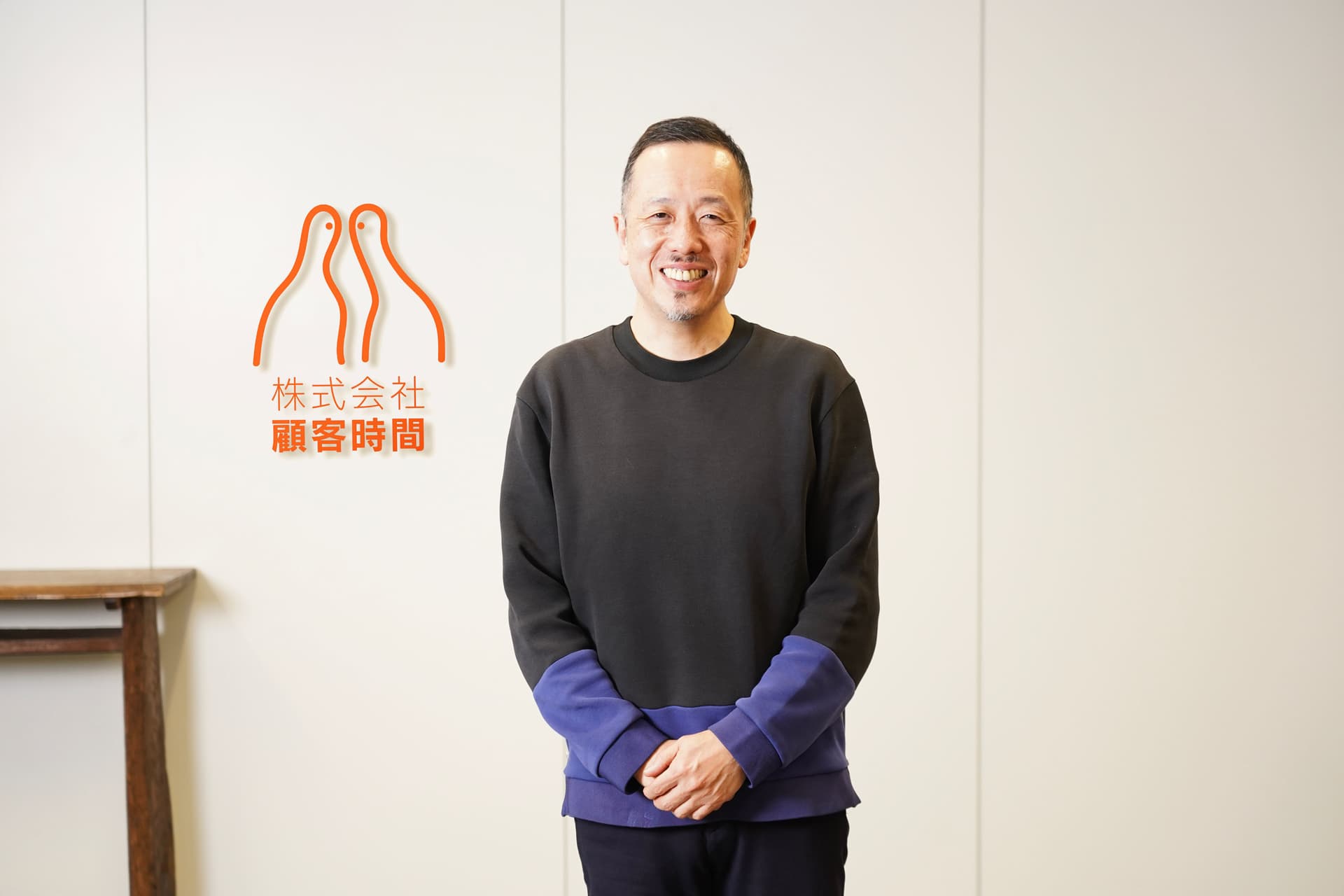 黒と青のシャツを着た笑顔の男性が、オレンジ色の日本の企業ロゴが書かれた白い壁の前に立っている。
