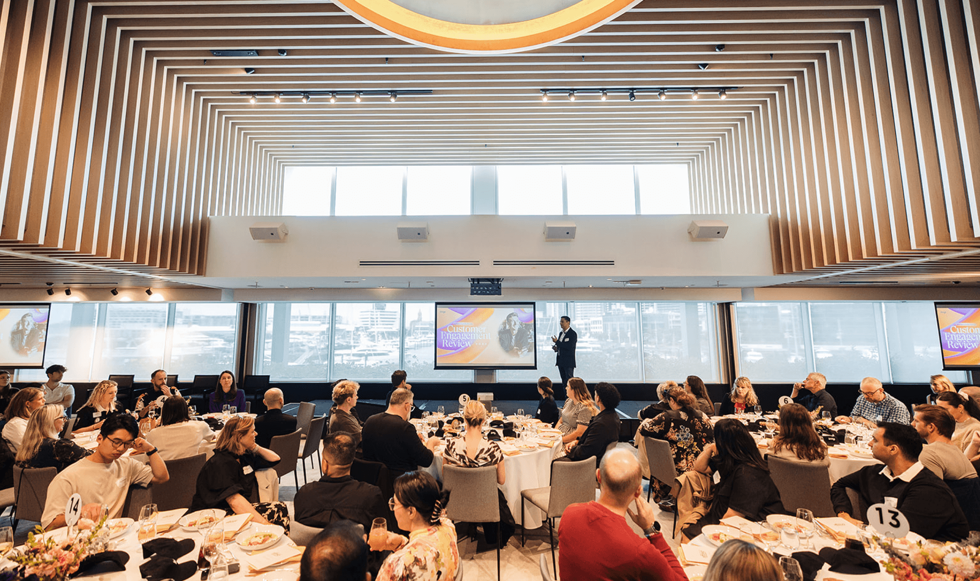 A speaker presents to an audience seated at round tables in a modern event hall with large screens.