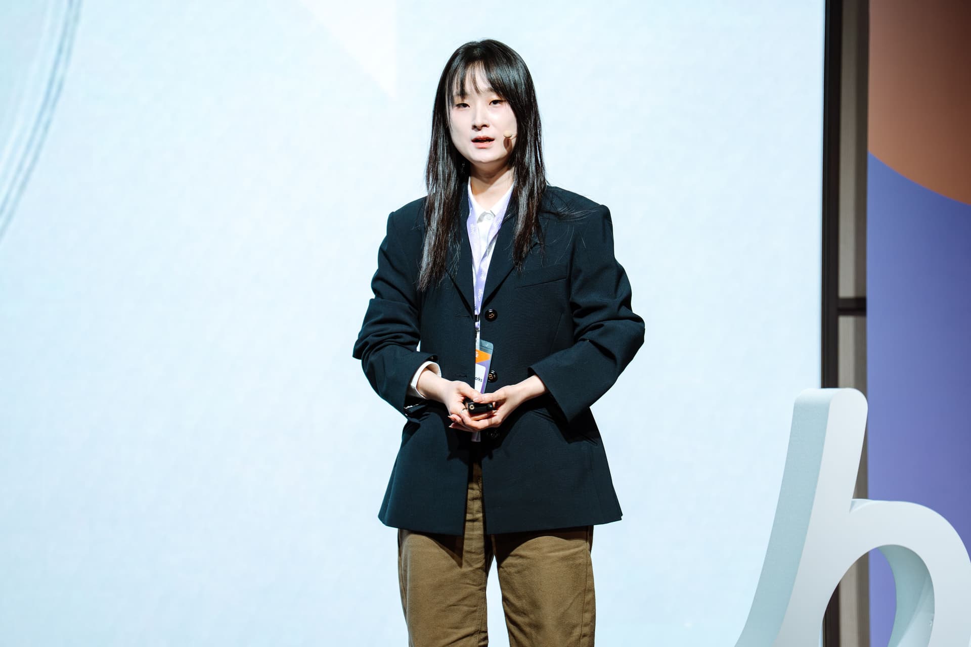 A young East Asian woman with long dark hair, wearing a blazer, stands on stage speaking into a microphone, hands clasped.