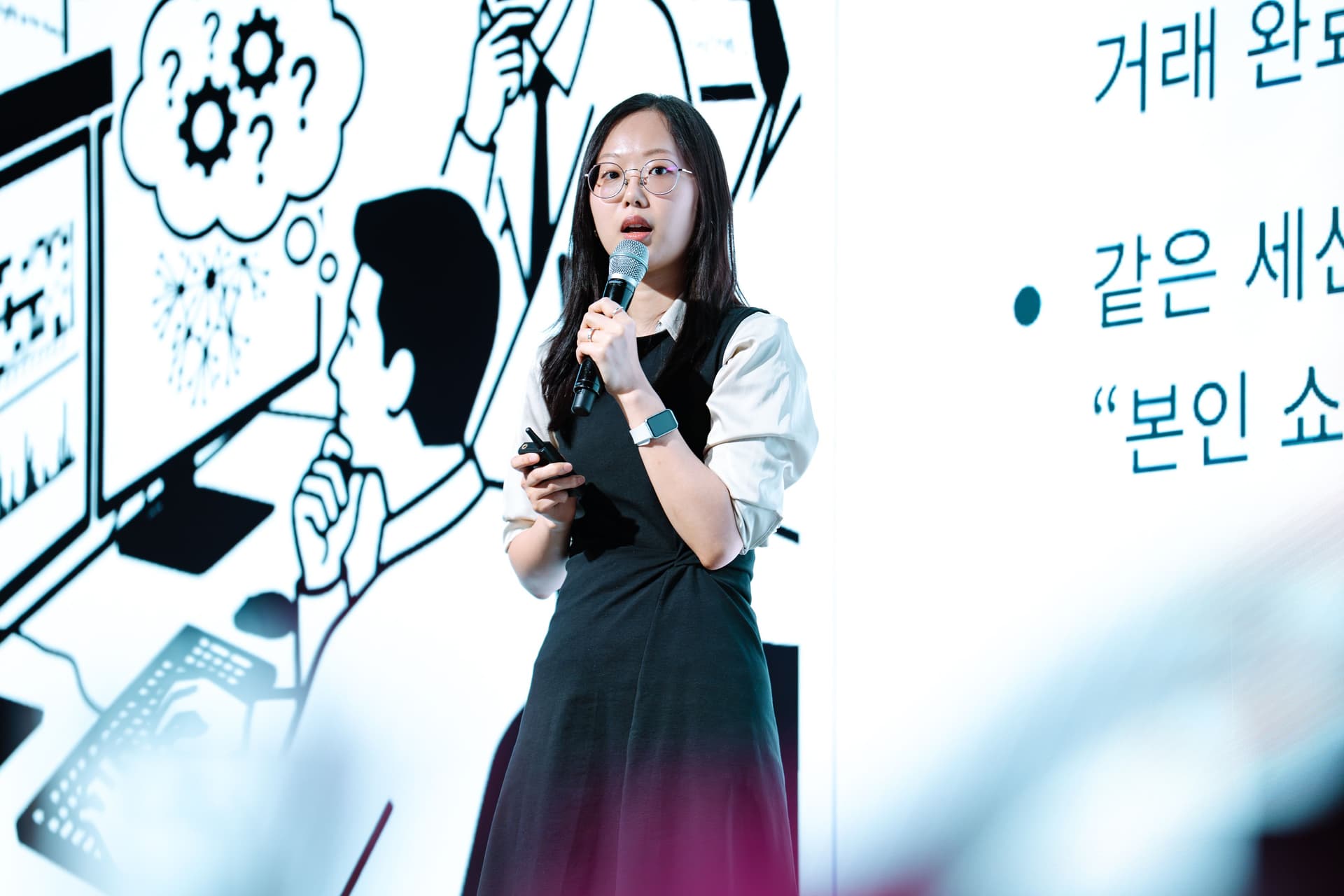 A woman speaks into a microphone in front of a screen displaying black and white illustrations of a person at a computer, charts, and Korean text.