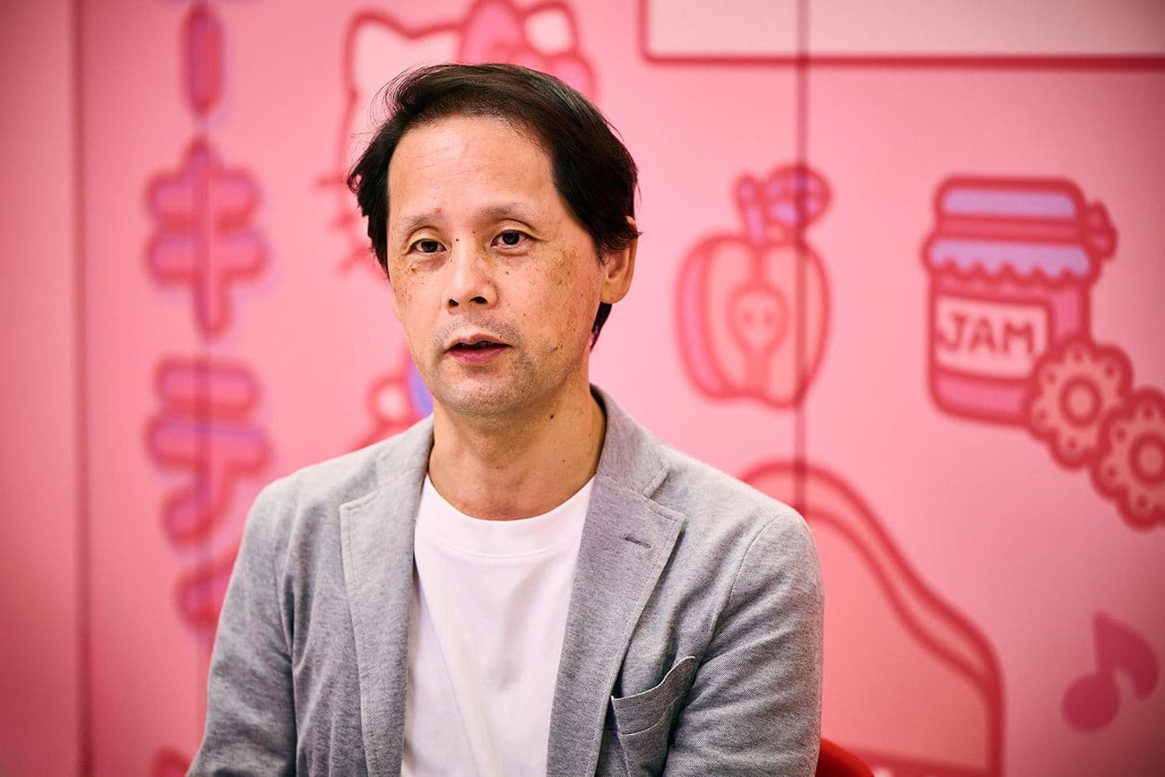 灰色のジャケットを着た男性(サンリオ デジタル事業本部 デジタル事業開発部 CX推進課 髙松 貴宏氏)が、ハローキティやその他の漫画のイラストで飾られたピンクの壁の前に座っている。