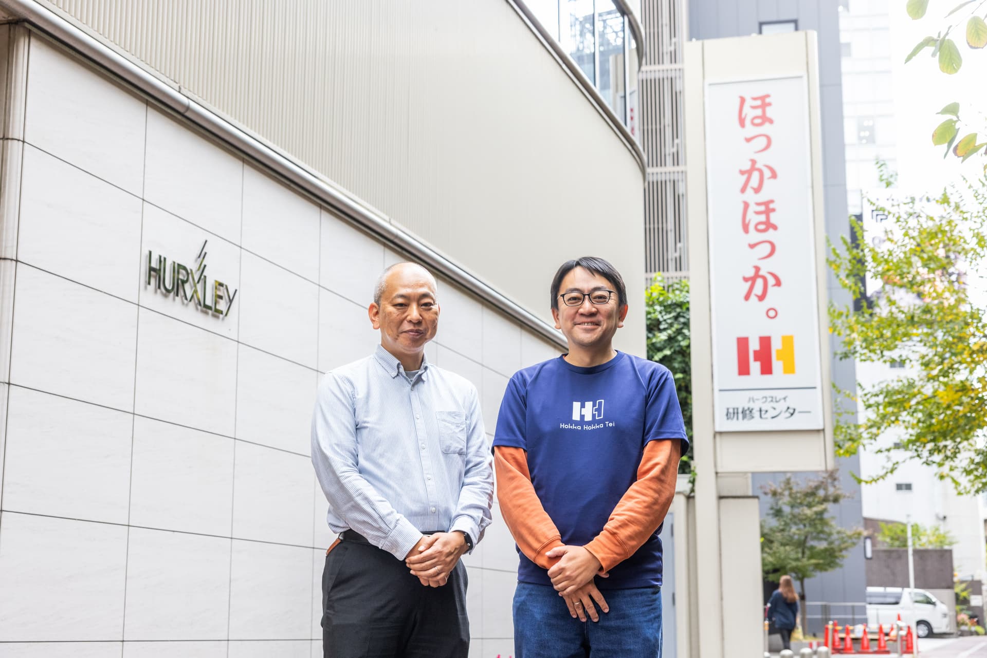 二人の男性が「ほっかほっか」と日本語の看板がある建物の外に立っている。