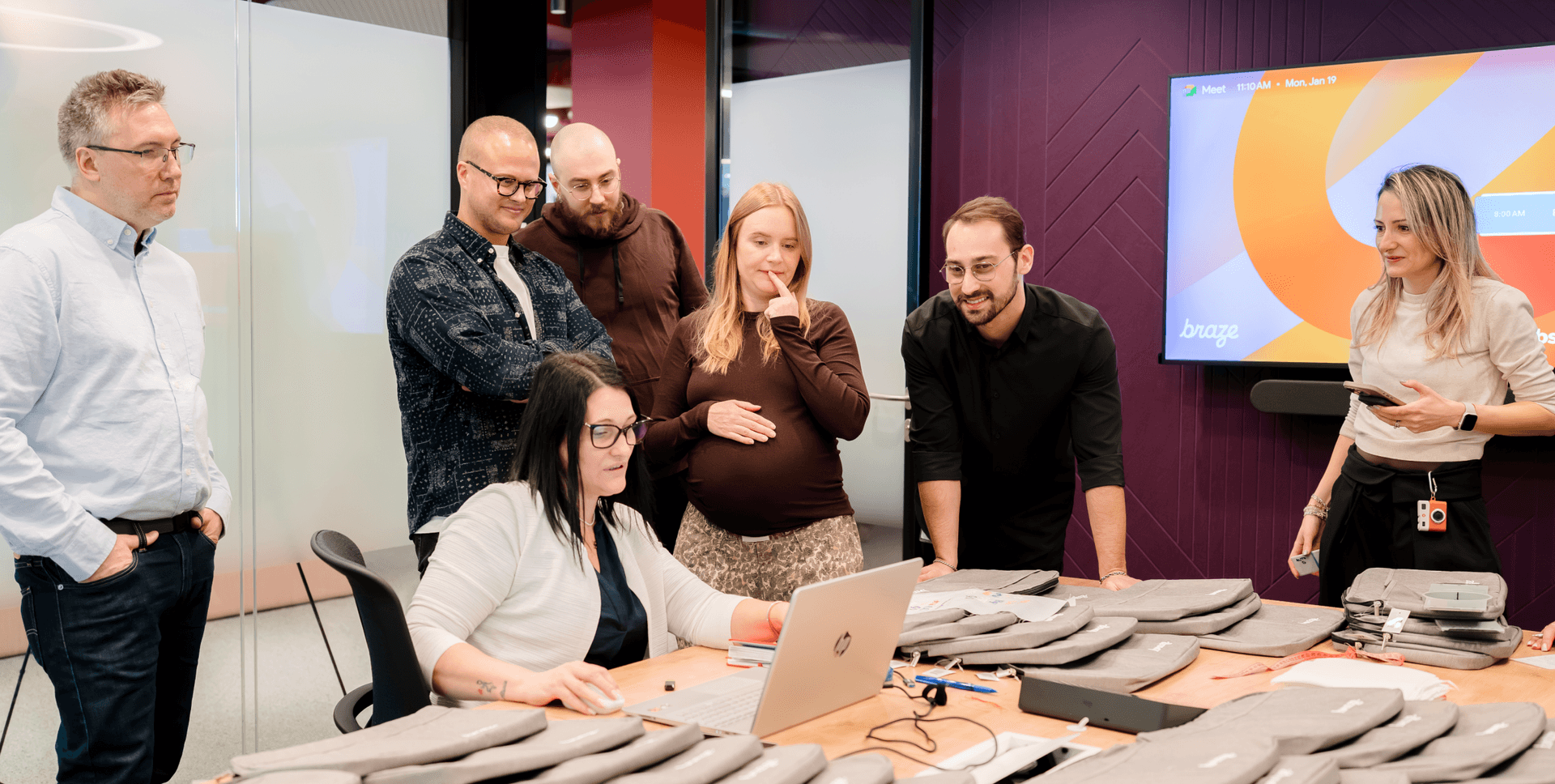 A group of seven people collaborating around a laptop in a modern office, with tech accessories on the table.