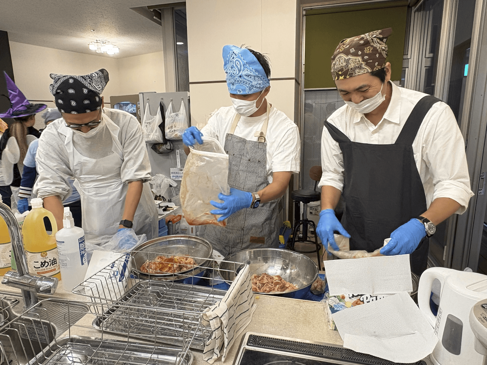 Three people in bandanas, masks, aprons, and blue gloves prepare food in a kitchen; one holds a bag of marinated meat.