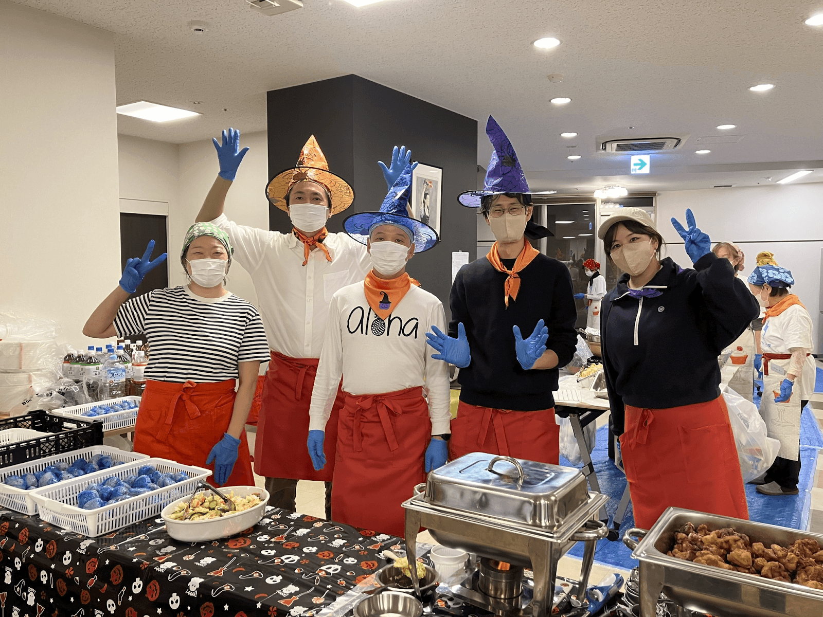 Six people in witches hats, masks, and blue gloves stand behind a Halloween-themed food table, gesturing hello.
