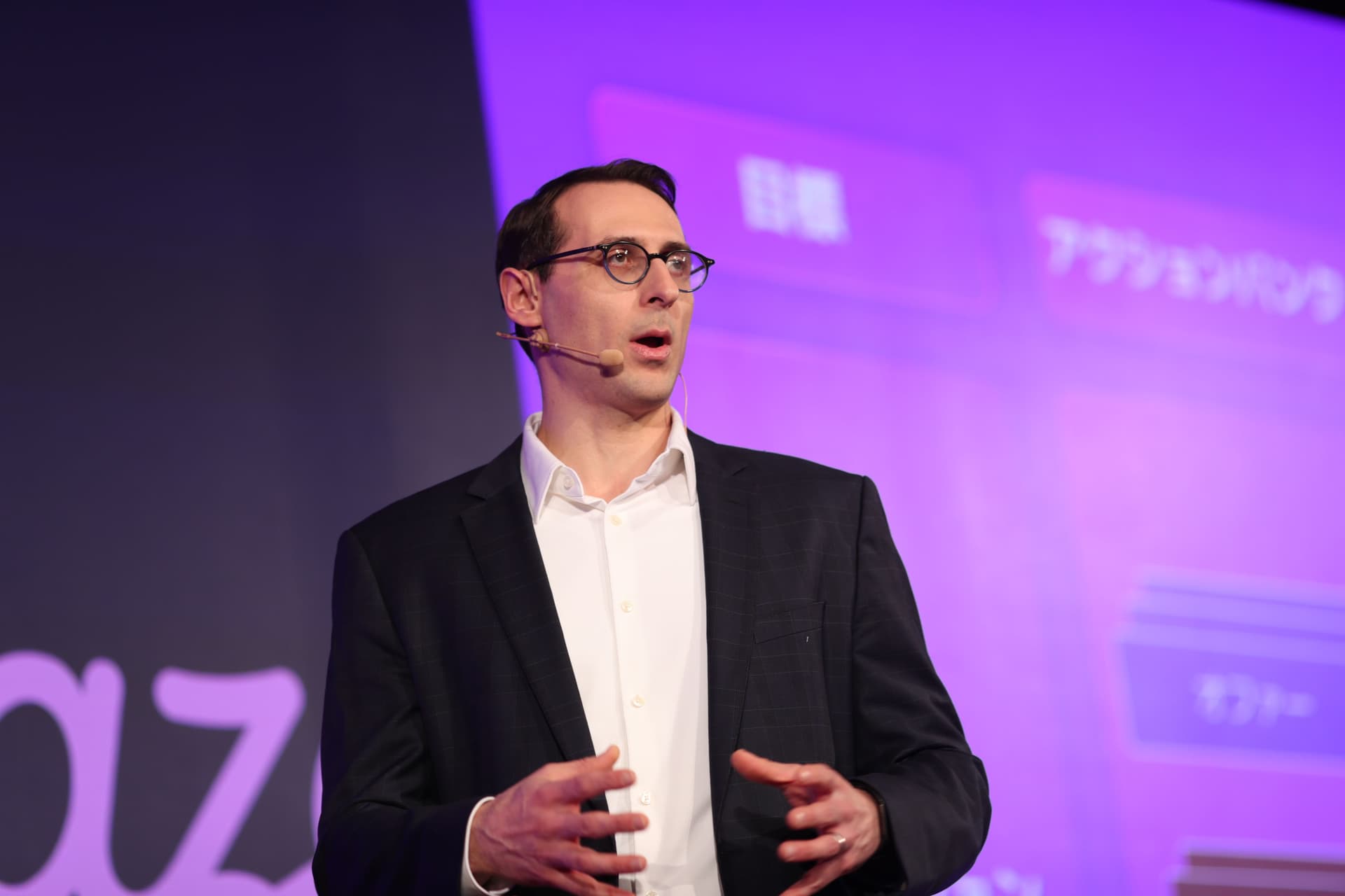 A man in a suit and glasses speaks into an ear microphone, gesturing on a purple-lit stage.