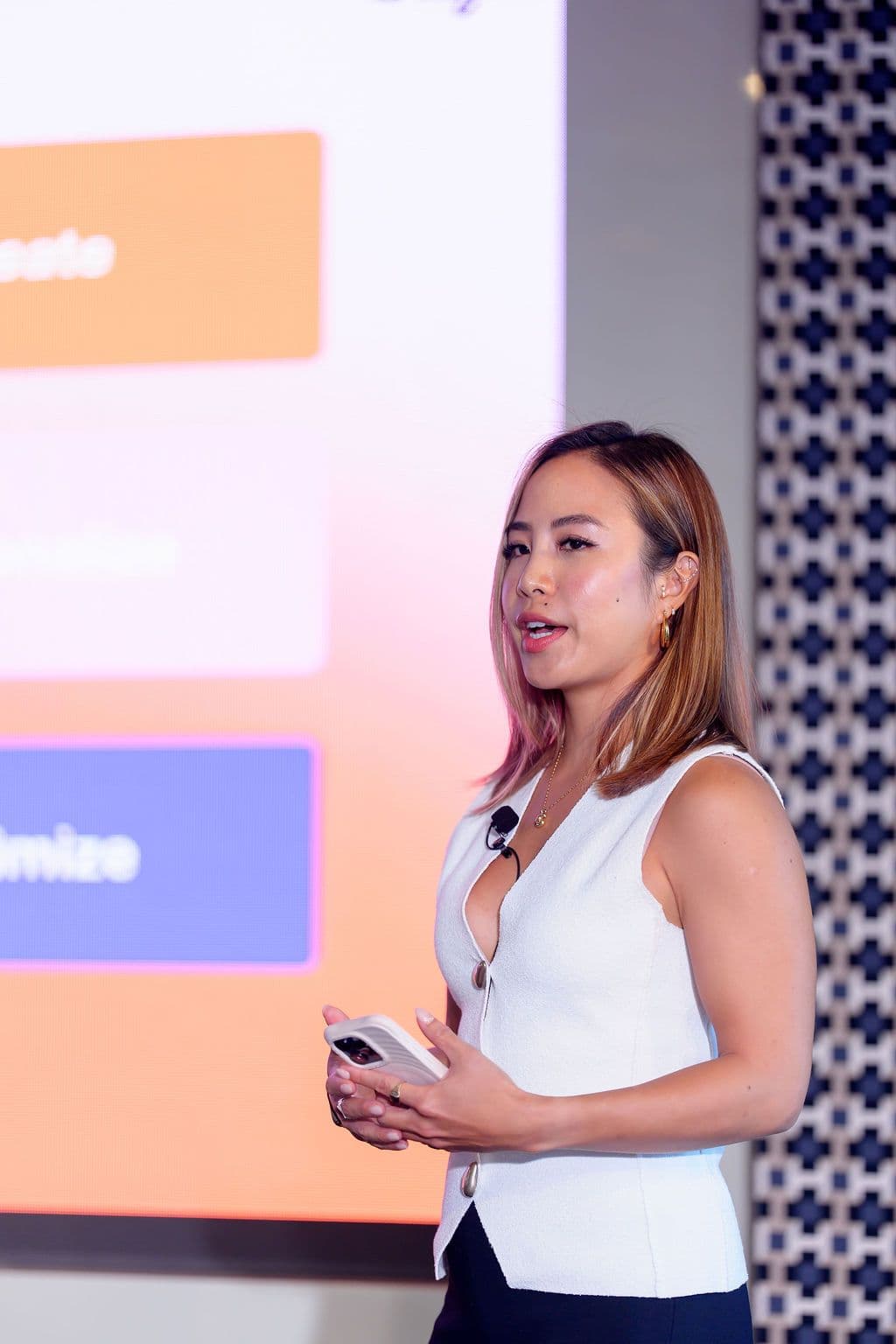 A woman with highlighted hair, wearing a white top and lavalier mic, speaks while holding a phone in front of a presentation screen.