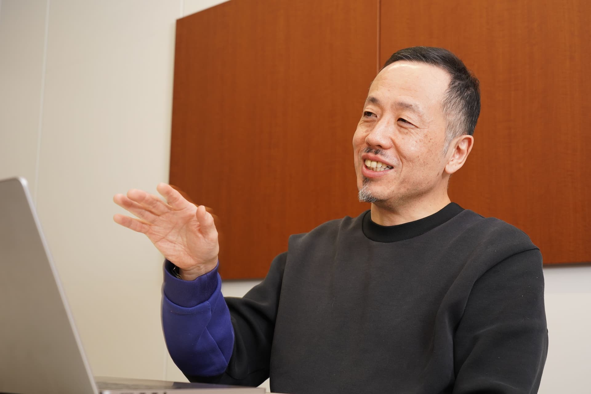 あごひげを生やした笑顔の中年アジア人男性が、ノートパソコンを置いたテーブルに座り、手でジェスチャーをしている。