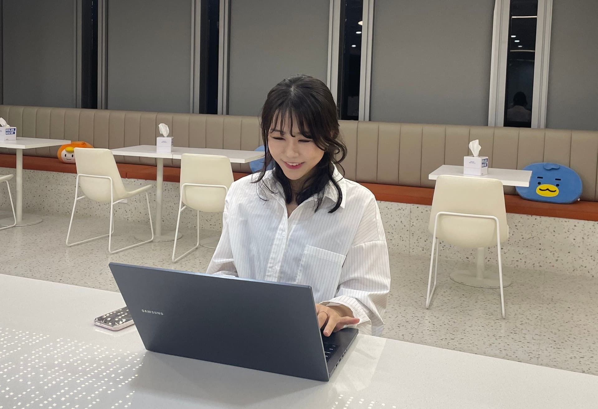 A smiling woman in a white shirt works on a laptop in a modern cafe.