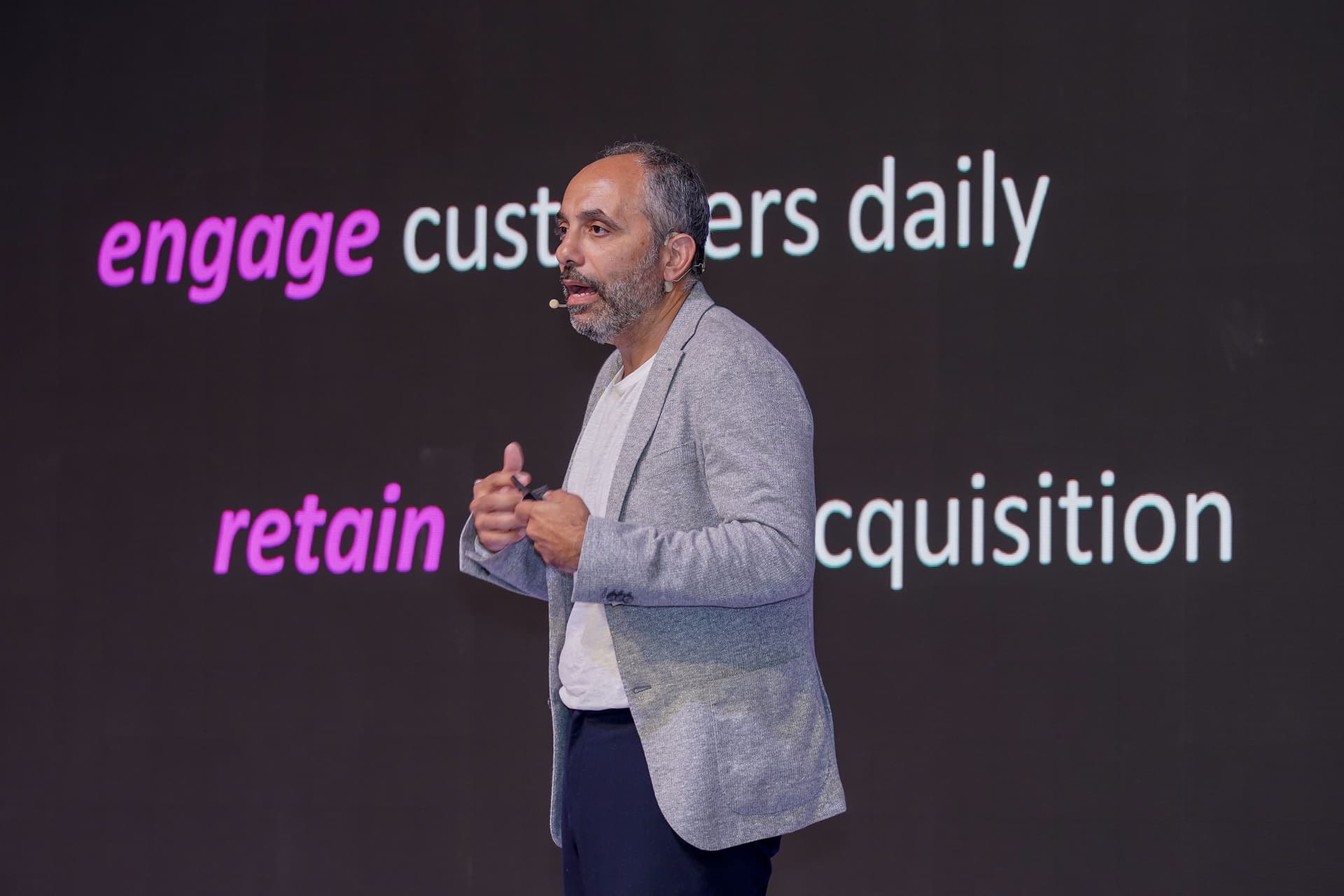 Man speaking on stage in front of a screen displaying "engage customers daily" and "retain acquisition."