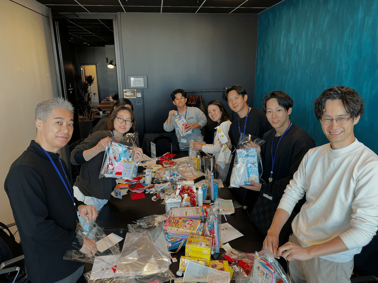笑顔の社員がテーブルを囲み、おもちゃやトイレタリー用品など様々な品々を透明な袋に詰めている。