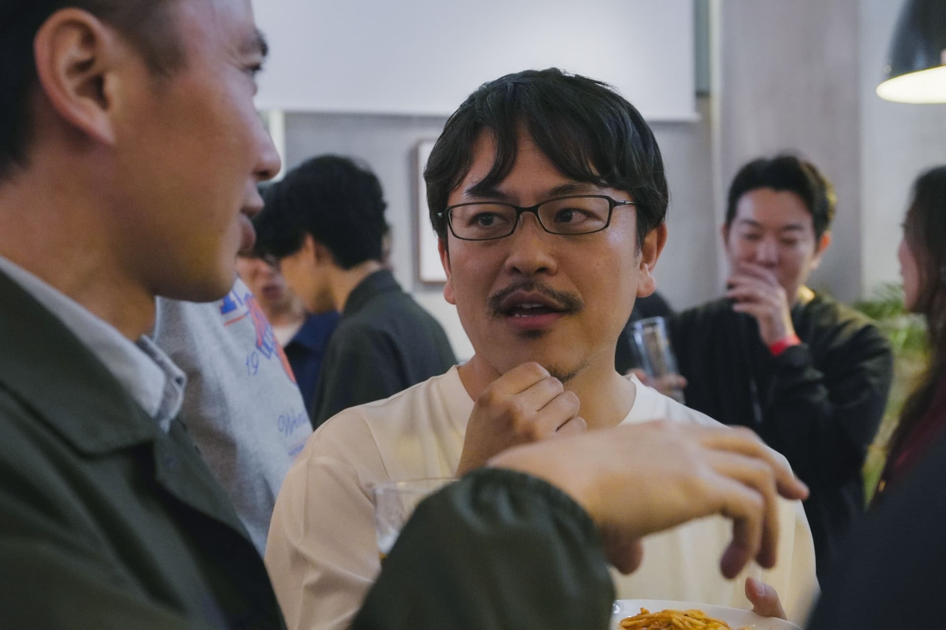 a man with glasses and a mustache is talking to another man at a party .