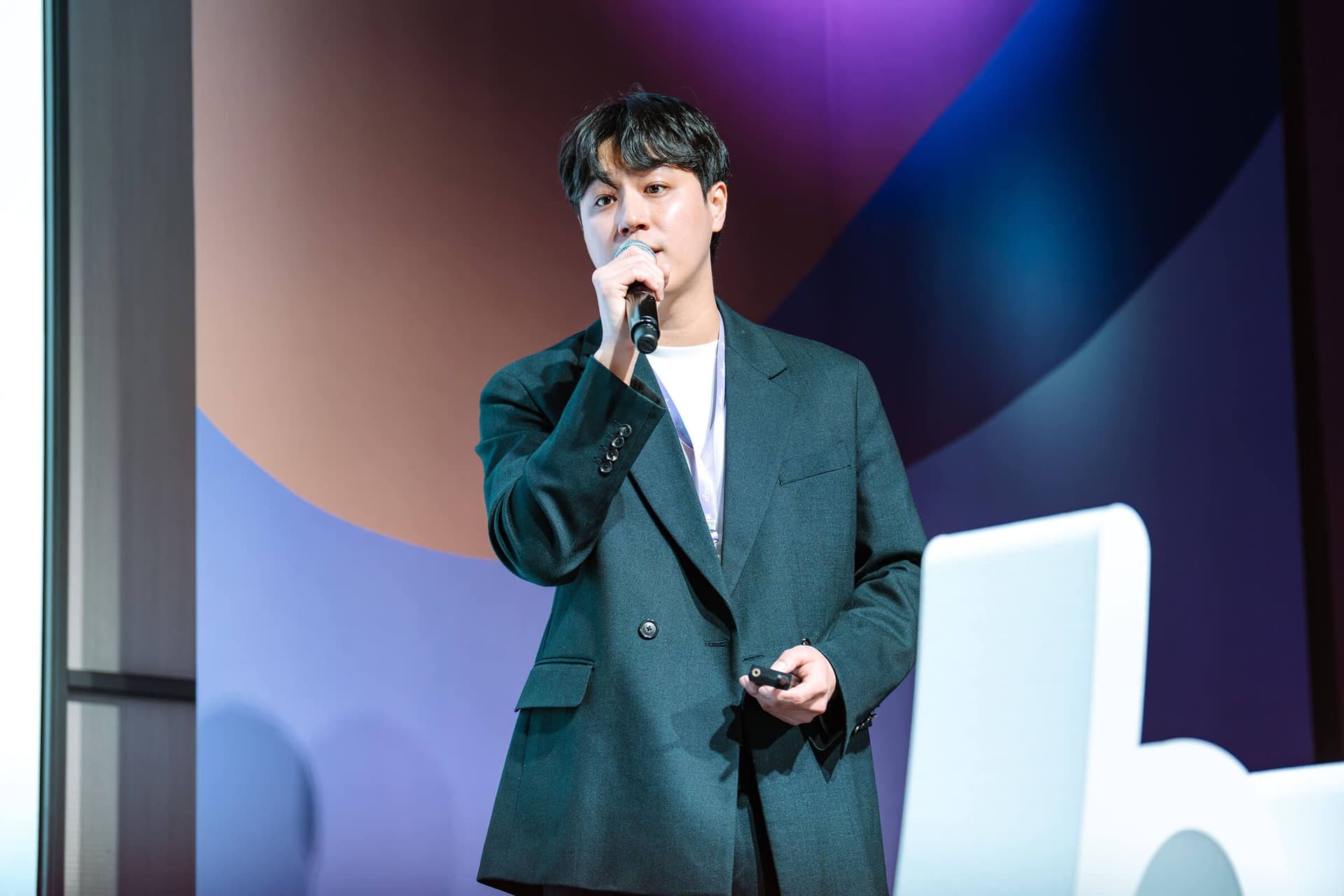 A speaker in a dark suit holds a microphone and clicker on a stage with a colorful backdrop.