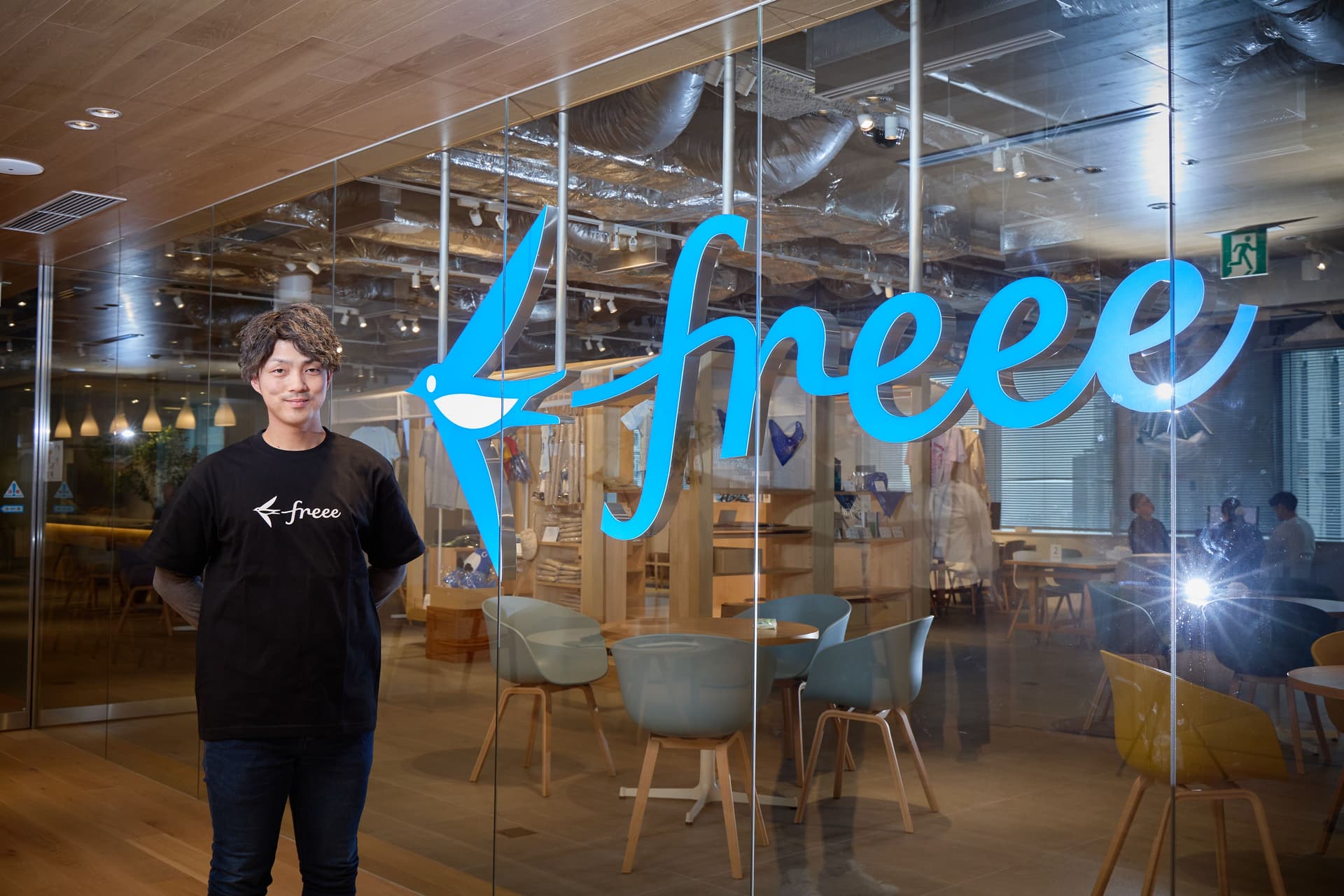黒い「freee」のTシャツを着た男性が、大きな青い「freee」のロゴが入ったガラス壁の前で微笑み、その向こうにはオフィスの内部が見えている。