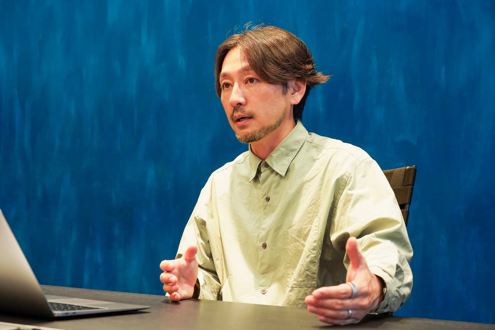 Man with shoulder-length hair and goatee in a light green shirt, gesturing while seated at a desk with a laptop against a blue wall.