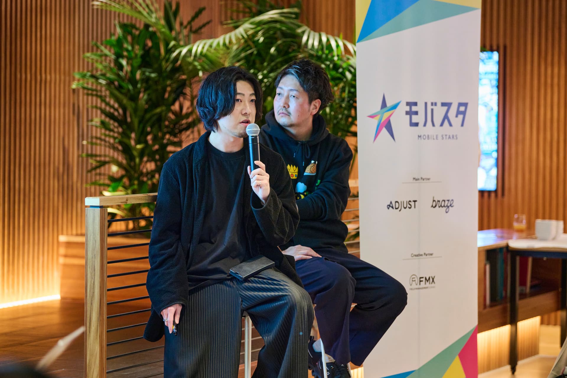 two men are sitting next to each other in front of a sign . one of the men is holding a microphone .