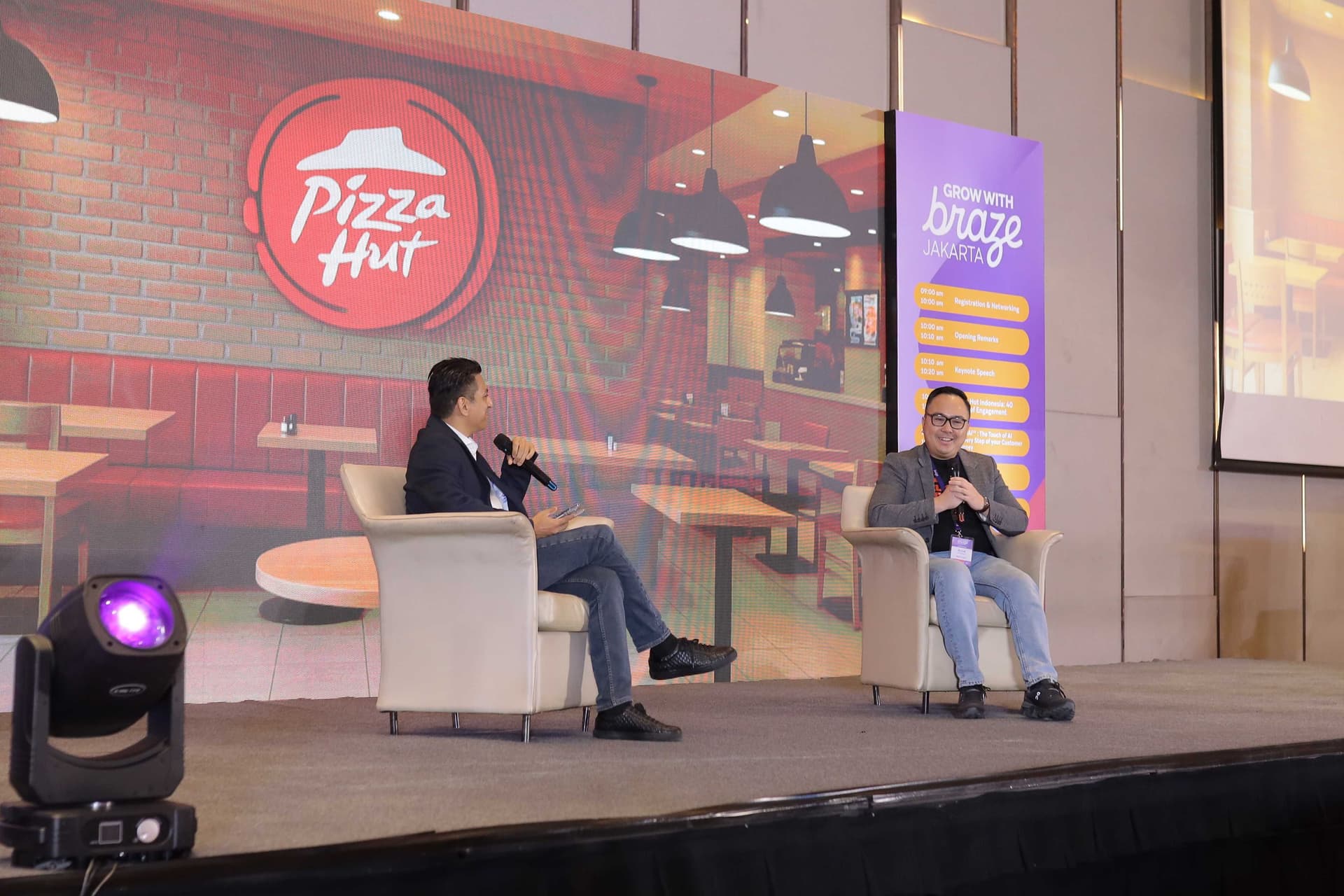two men are sitting in chairs on a stage in front of a pizza hut restaurant .