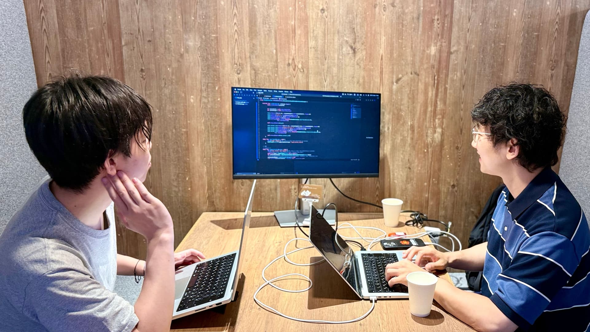 two men are sitting at a table with laptops and a computer .