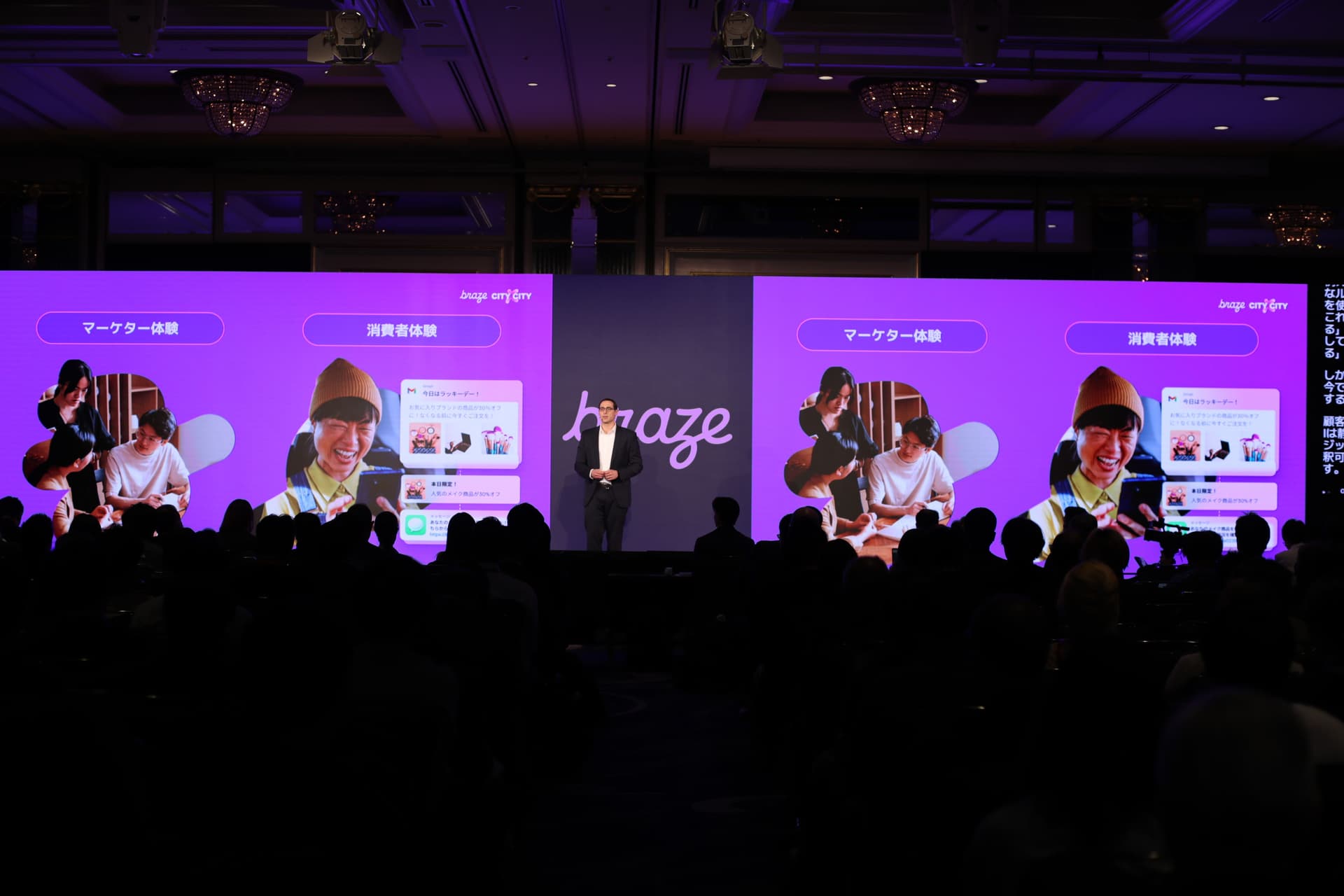 A presenter on stage next to a large screen displaying the Braze logo, purple graphics of people using devices and Japanese text, facing an audience.