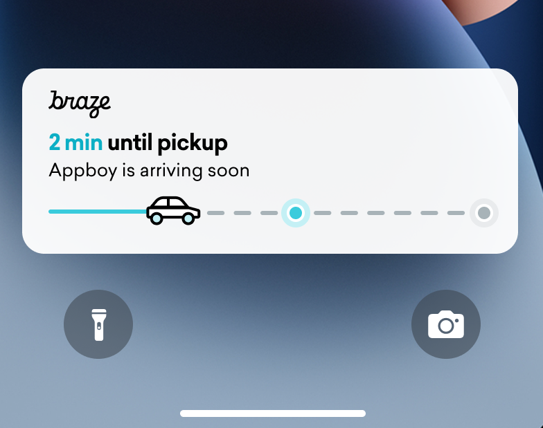 Atividade ao vivo de um rastreador de entregas em uma tela de bloqueio do iPhone. Uma barra de status com um carro está quase totalmente preenchida. O texto diz "2 min until pickup" (2 minutos até a coleta)